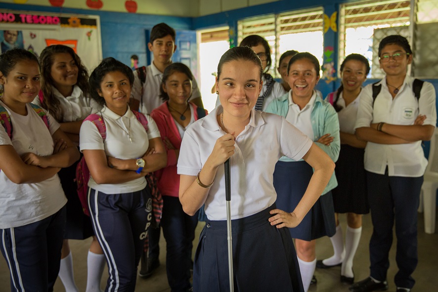 A group of students stand in a classroom. The girl in front is smiling and has one hand on her hip and holds a white cane in her other hand
